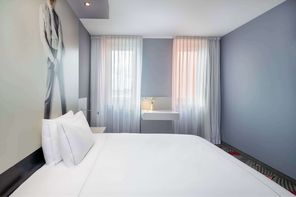 Una habitación de hotel moderna con una cama grande, una mesita de noche, un ventanal con cortinas translúcidas y un estante en la pared con un jarrón.