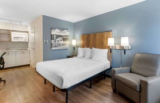 Habitación de hotel con cama, una silla, escritorio con lámpara, microondas y un cuadro en la pared.