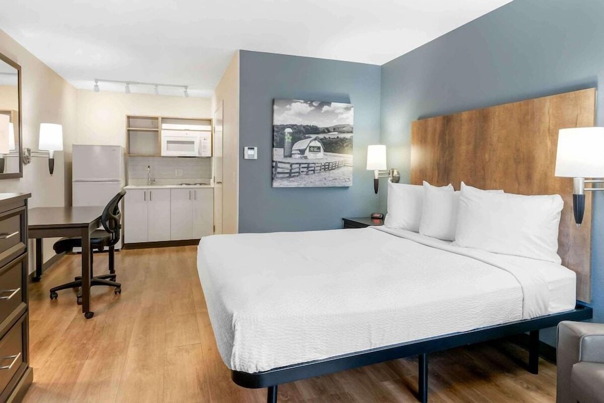 A hotel room with a bed, desk, chair, kitchenette, and artwork on the wall.