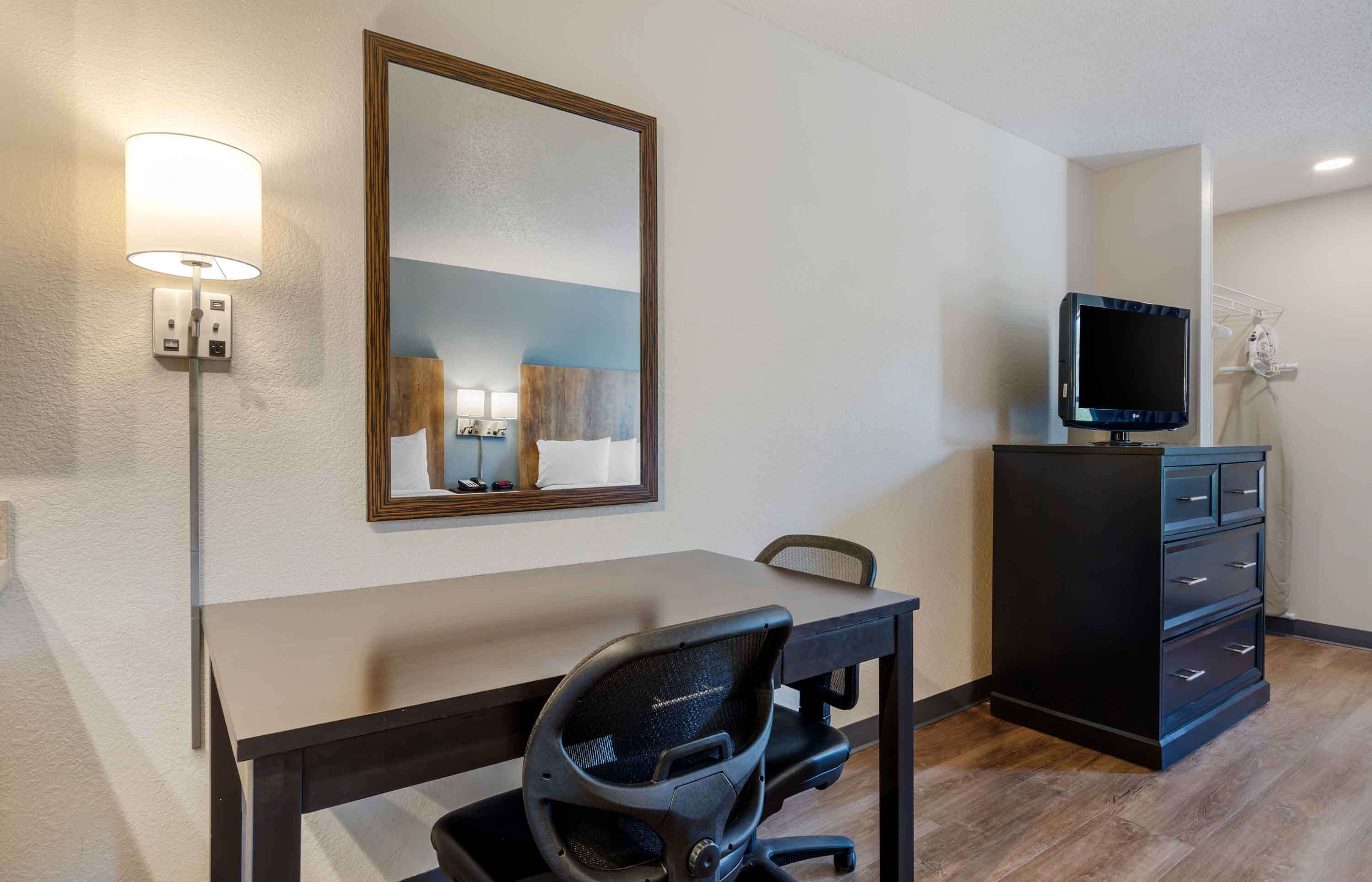 A hotel room with a desk, chair, TV, dresser, and a mirror.
