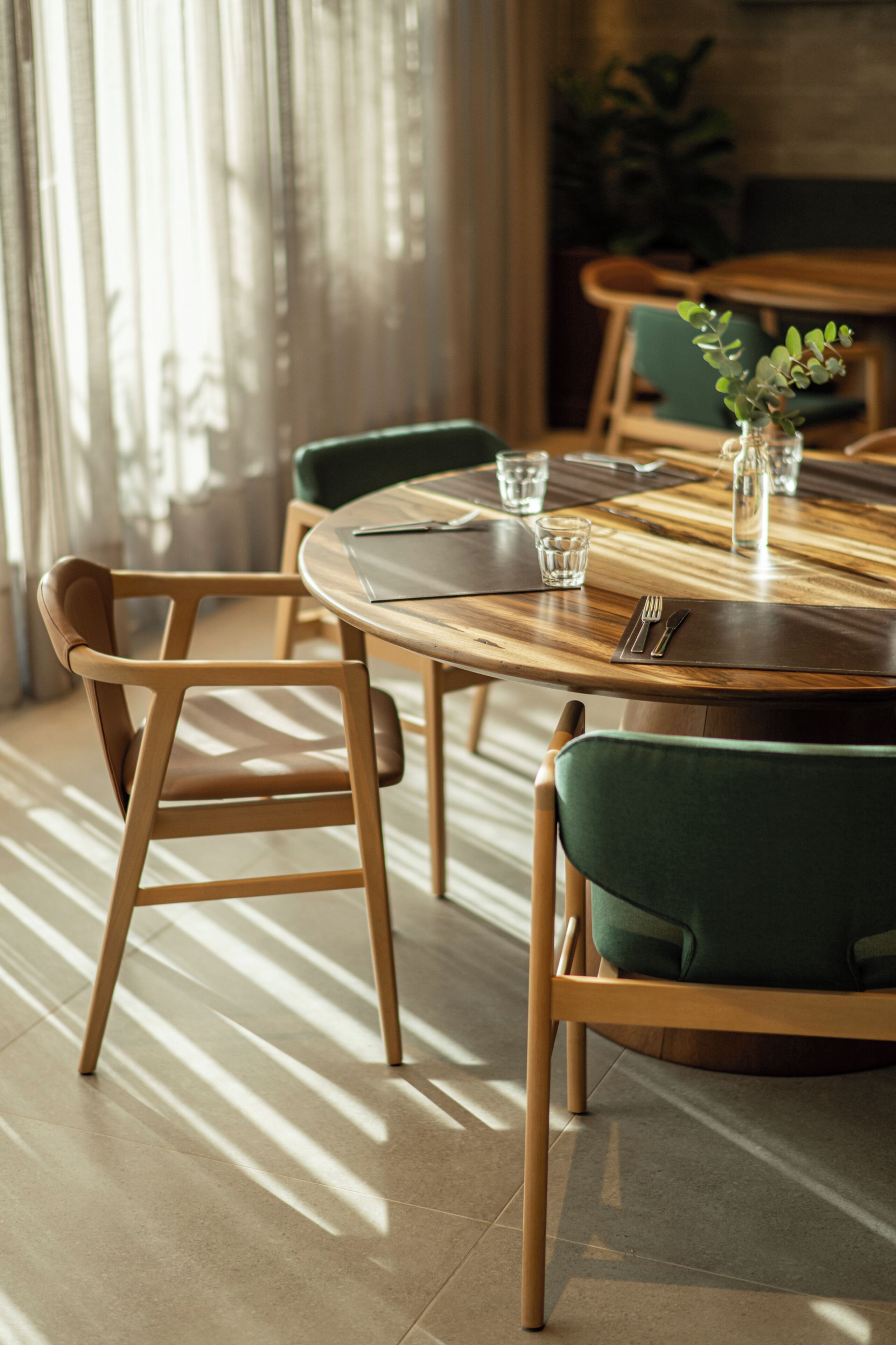 A dining area with a round wooden table, two green upholstered chairs, and a vase with a plant.