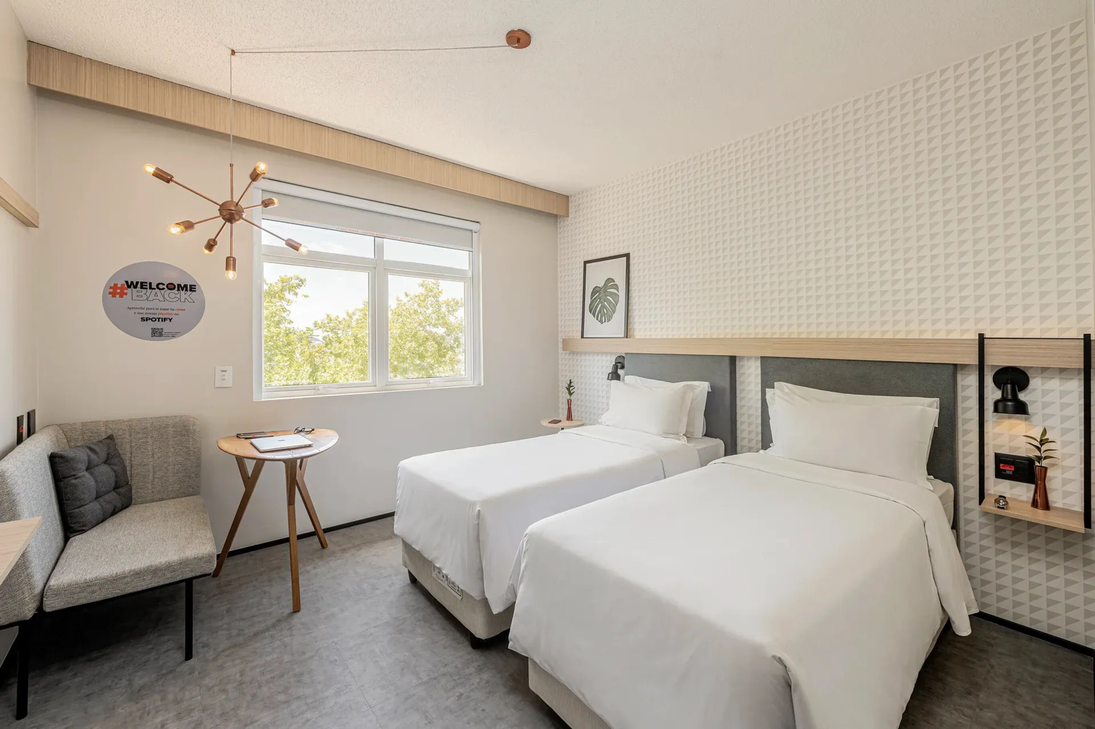 A modern hotel room with two beds, a sofa, a small table, a wall-mounted shelf, and a window with a view.