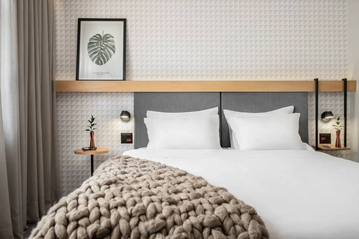 Ein Hotelzimmer mit einem großen Bett, einem gerahmten Kunstwerk mit Blättern, einem kleinen Tisch mit Pflanze und zwei Nachttischlampen.