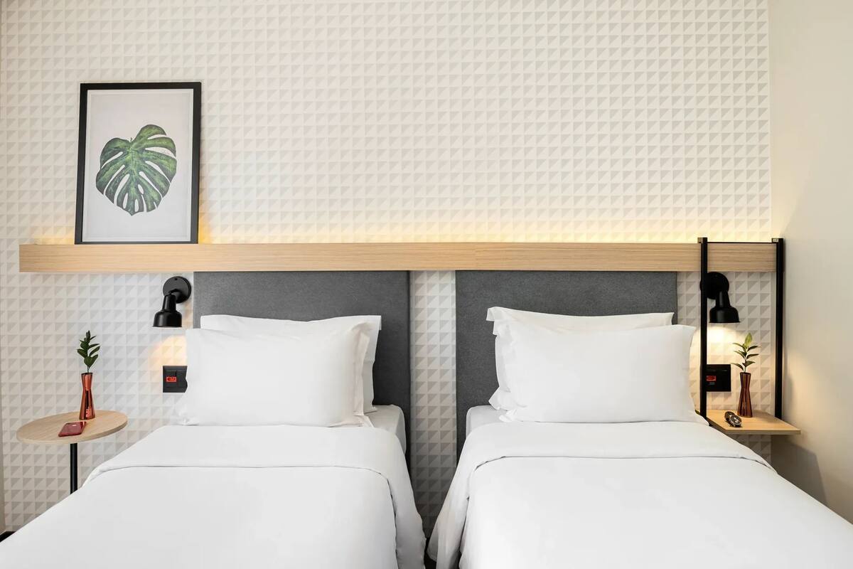 Ein Hotelzimmer mit zwei Betten, einer gerahmten Pflanze an der Wand und Nachttischen mit Lampen.