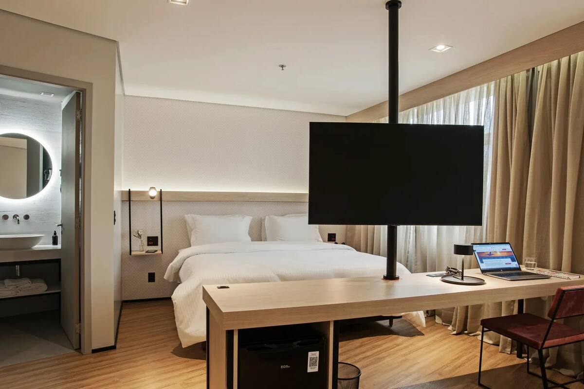 Ein modernes Hotelzimmer mit einem großen Bett, einem Schreibtisch mit einem Laptop, einem Fernseher und einem Badezimmer mit einem runden Spiegel und Waschbecken.