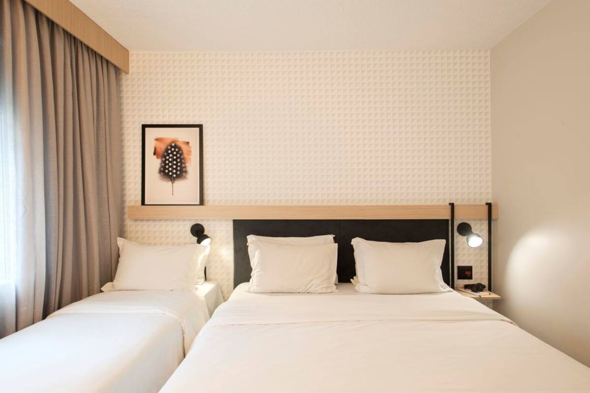 Ein Hotelzimmer mit zwei Betten, einem Kopfteil, einem gerahmten Kunstwerk, einem Nachttisch und einer Nachttischlampe.