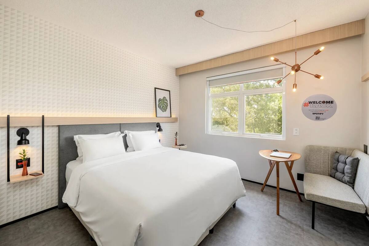 Ein modernes Hotelzimmer mit einem großen Bett, einem kleinen Tisch, einem Sessel, einem Fenster mit Aussicht und einem 'Welcome Back'-Schild.