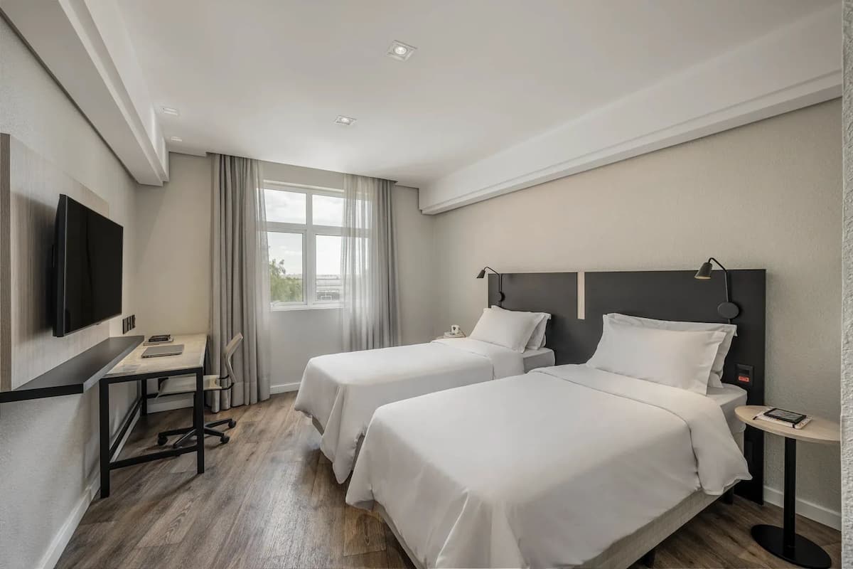 Ein Hotelzimmer mit zwei Betten, einem Schreibtisch, einem Fernseher und einem Fenster mit Vorhängen.