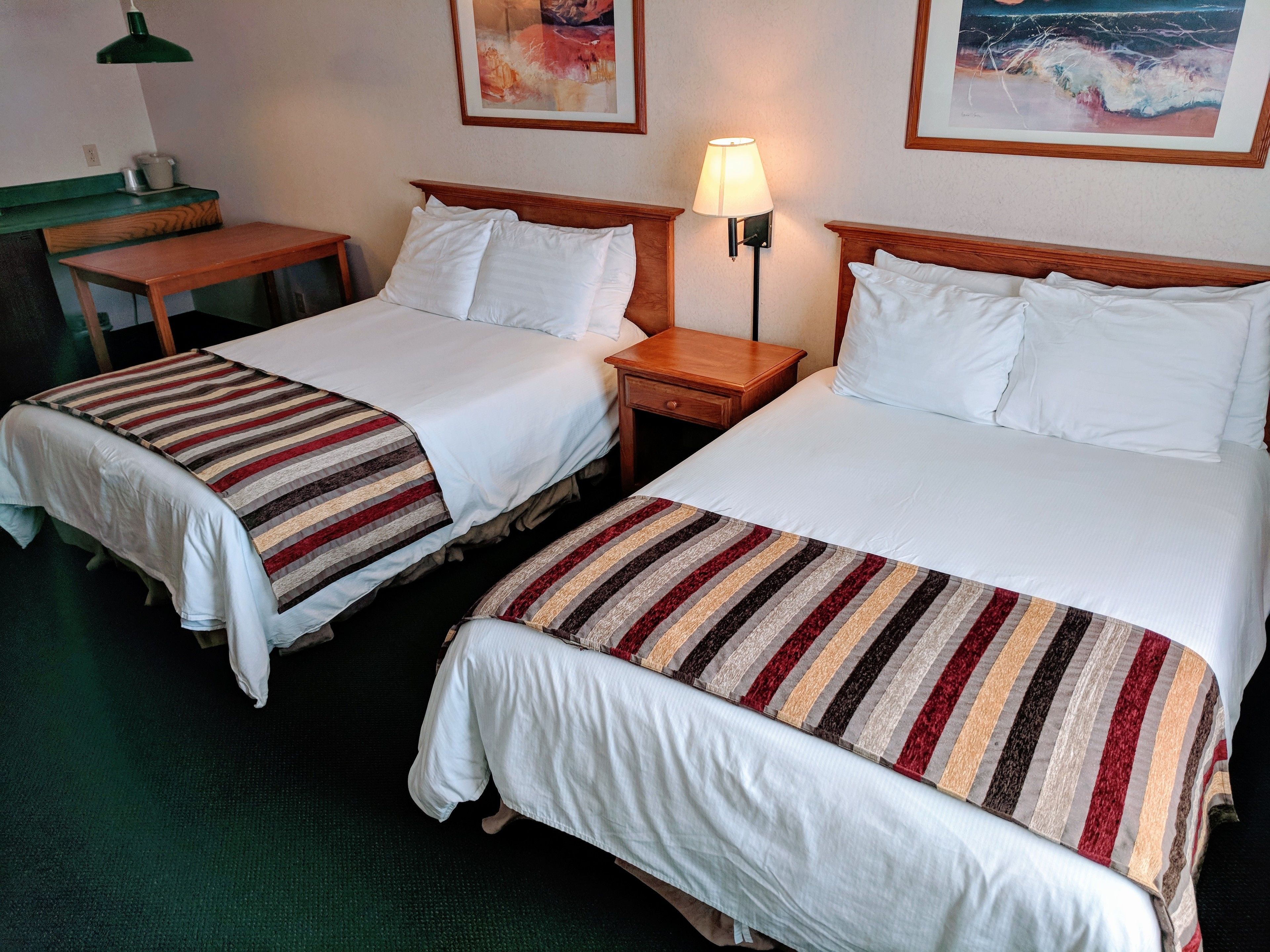 Une chambre d’hôtel avec deux lits, chacun recouvert d’une couverture à rayures et agrémenté de coussins blancs. On y trouve une petite table en bois et une lampe sur un des côtés.