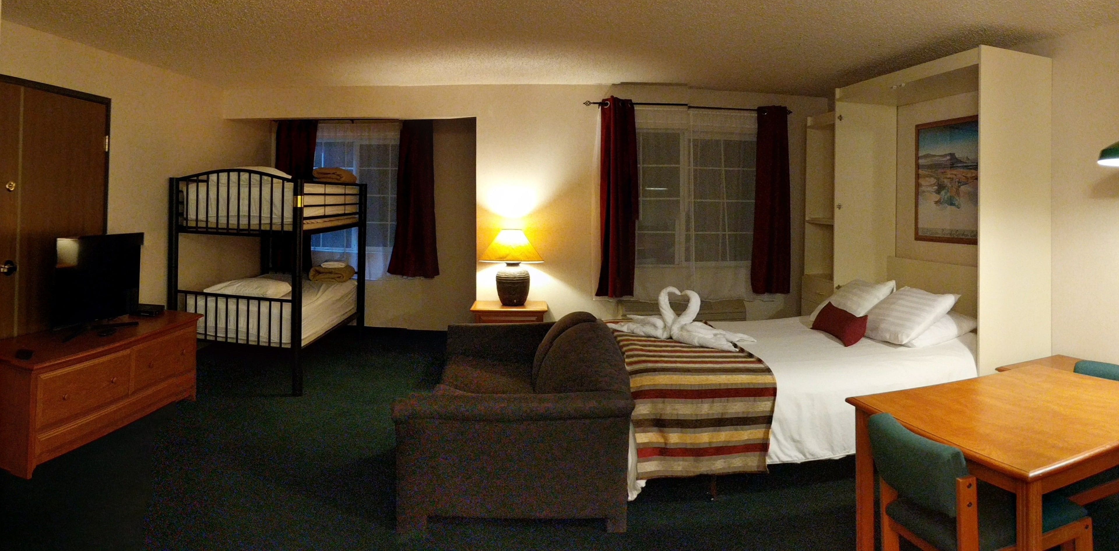A hotel room with bunk beds, a single bed, a desk, a chair, a lamp, and a painting on the wall.