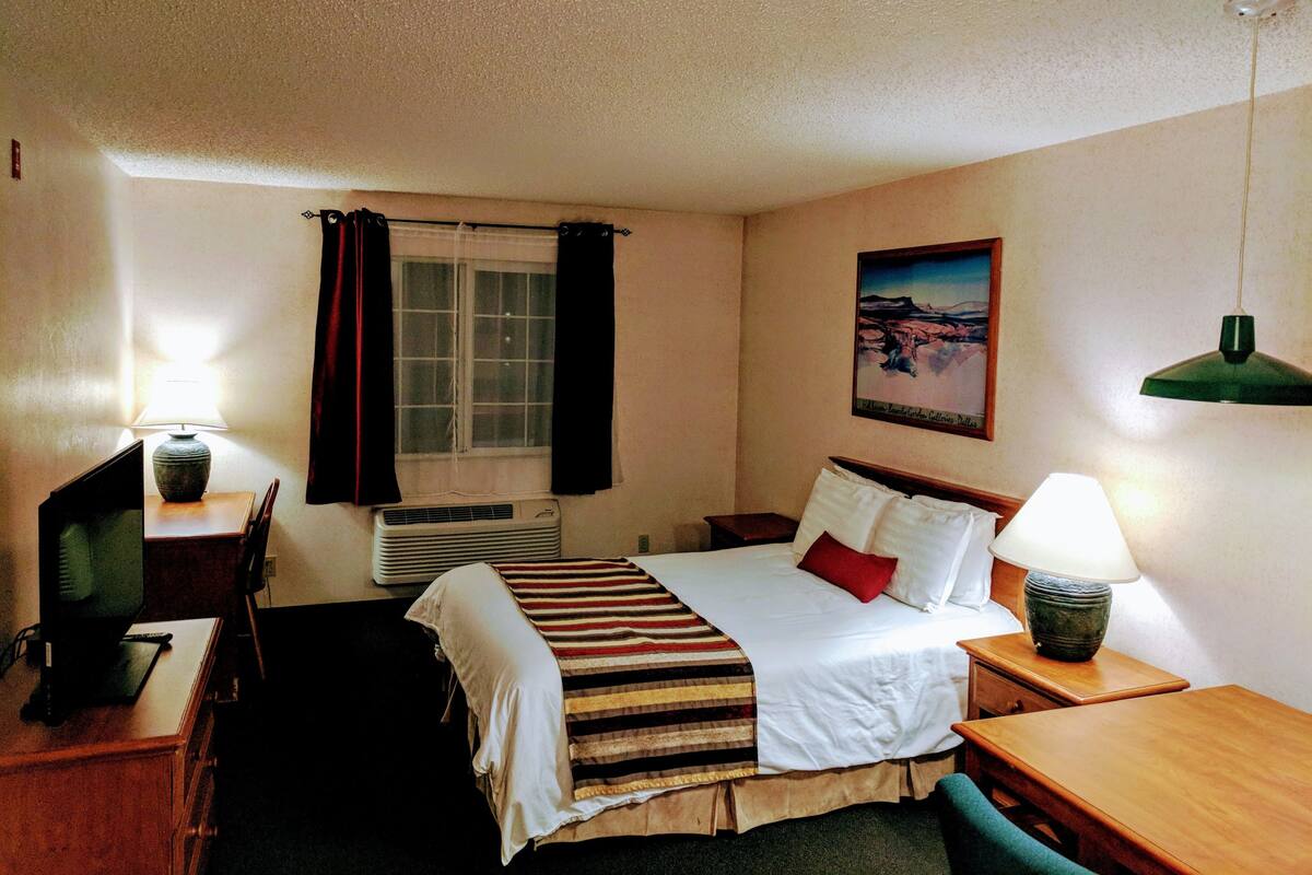 Ett hotellrum med en säng, ett skrivbord, en stol, en lampa och en tavla på väggen.