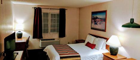 Una habitación de hotel con cama, escritorio, silla, lámpara y un cuadro en la pared.