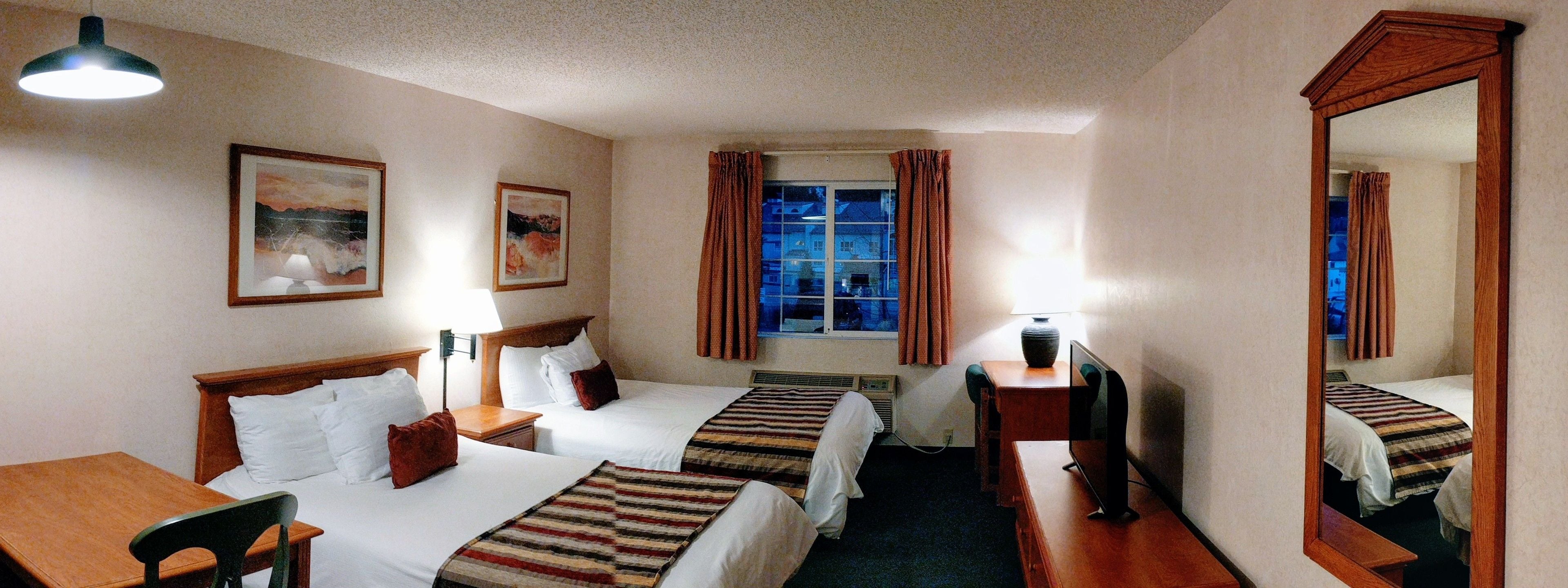 A hotel room with two beds, a desk, a chair, a TV, a window with curtains, and a mirror.