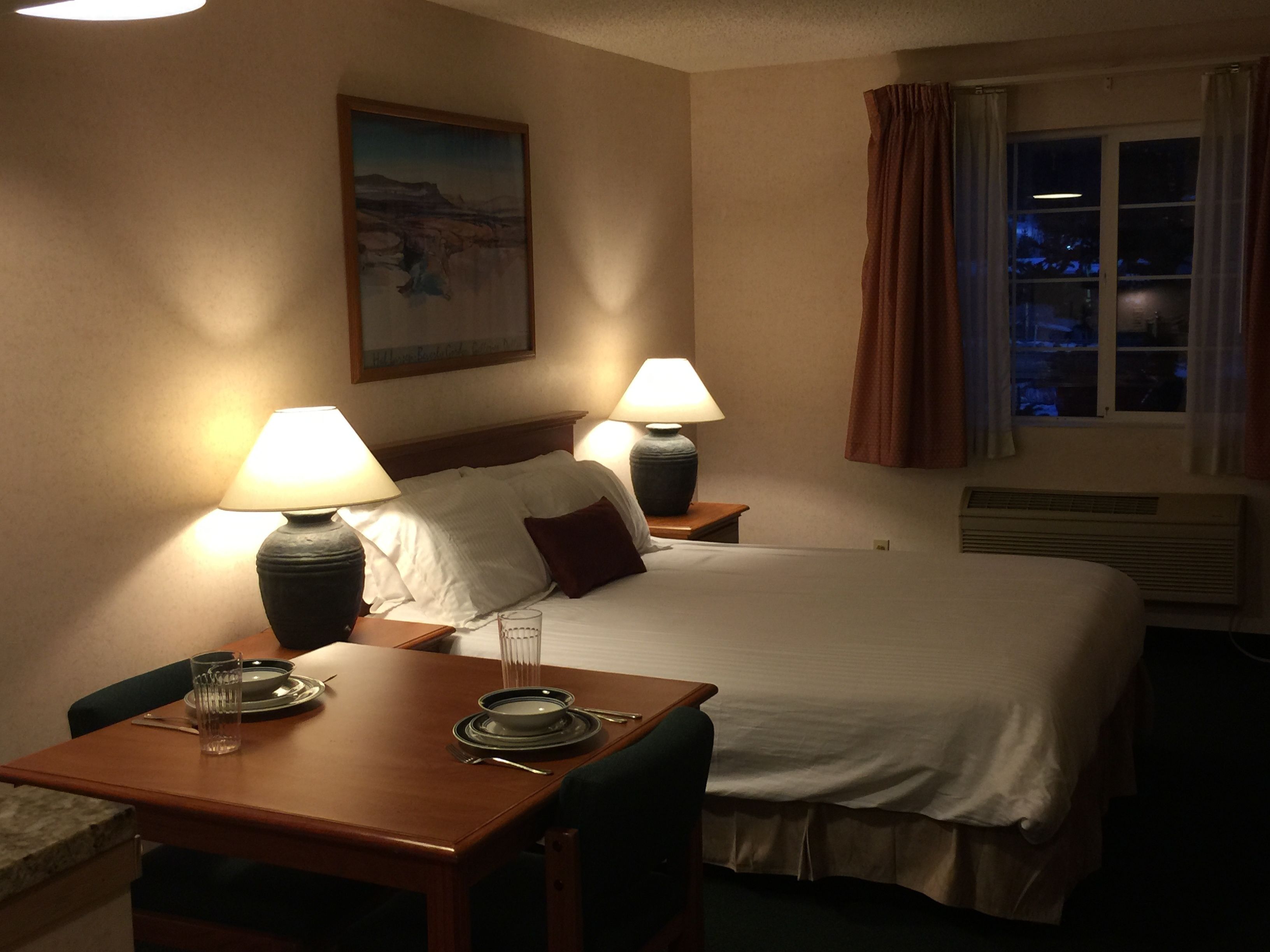 A hotel room with a bed, two bedside lamps, a dining table with plates and glasses, a chair, a painting on the wall, and a window with curtains.