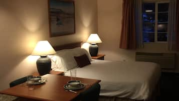 A hotel room with a bed, two bedside lamps, a dining table with plates and glasses, a chair, a painting on the wall, and a window with curtains.
