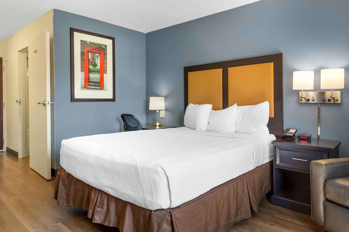 A hotel room with a large bed, a nightstand with a lamp, a chair, and a framed picture on the wall.