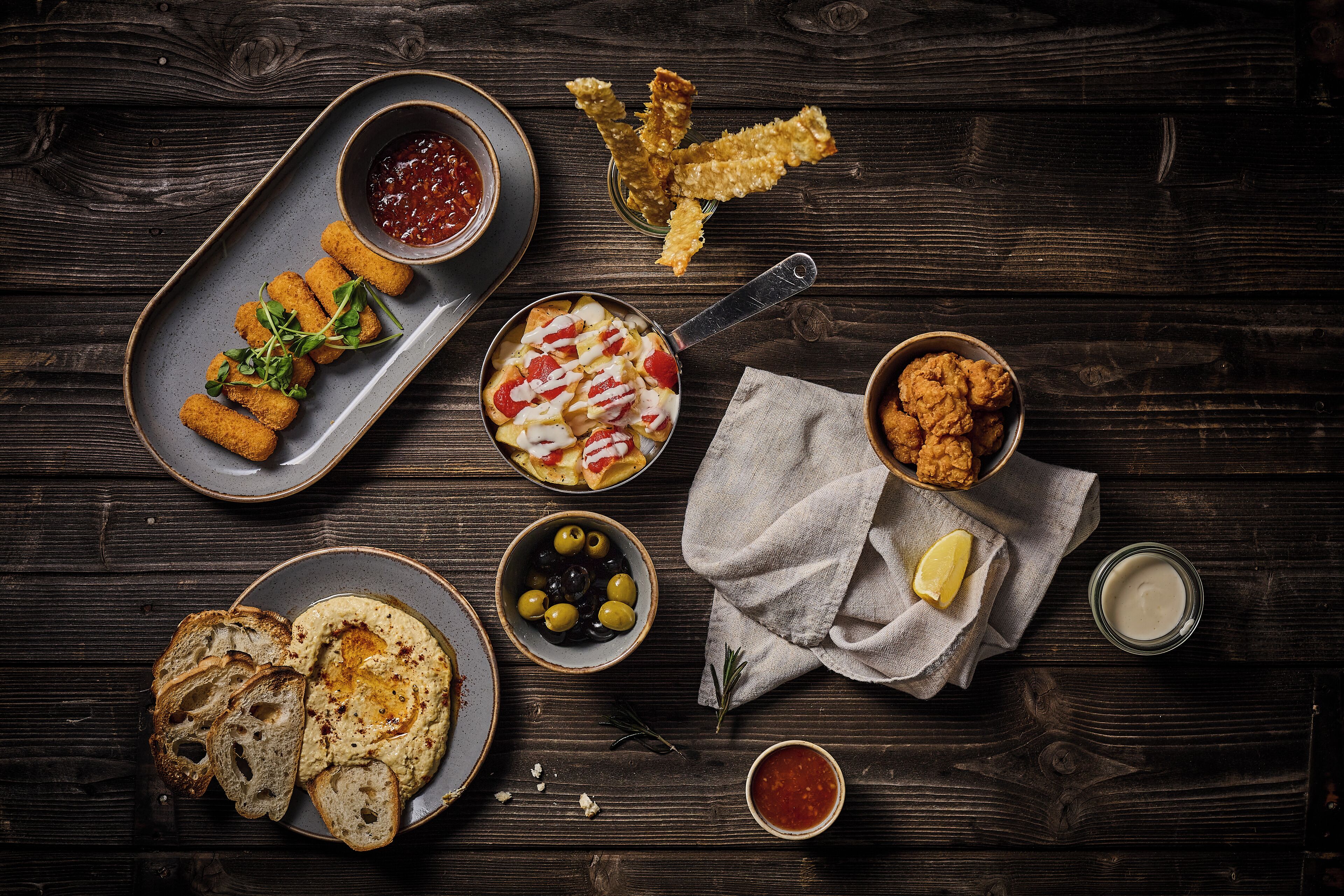 Diversos alimentos como pão, homus, azeitonas e petiscos fritos sobre uma mesa de madeira.