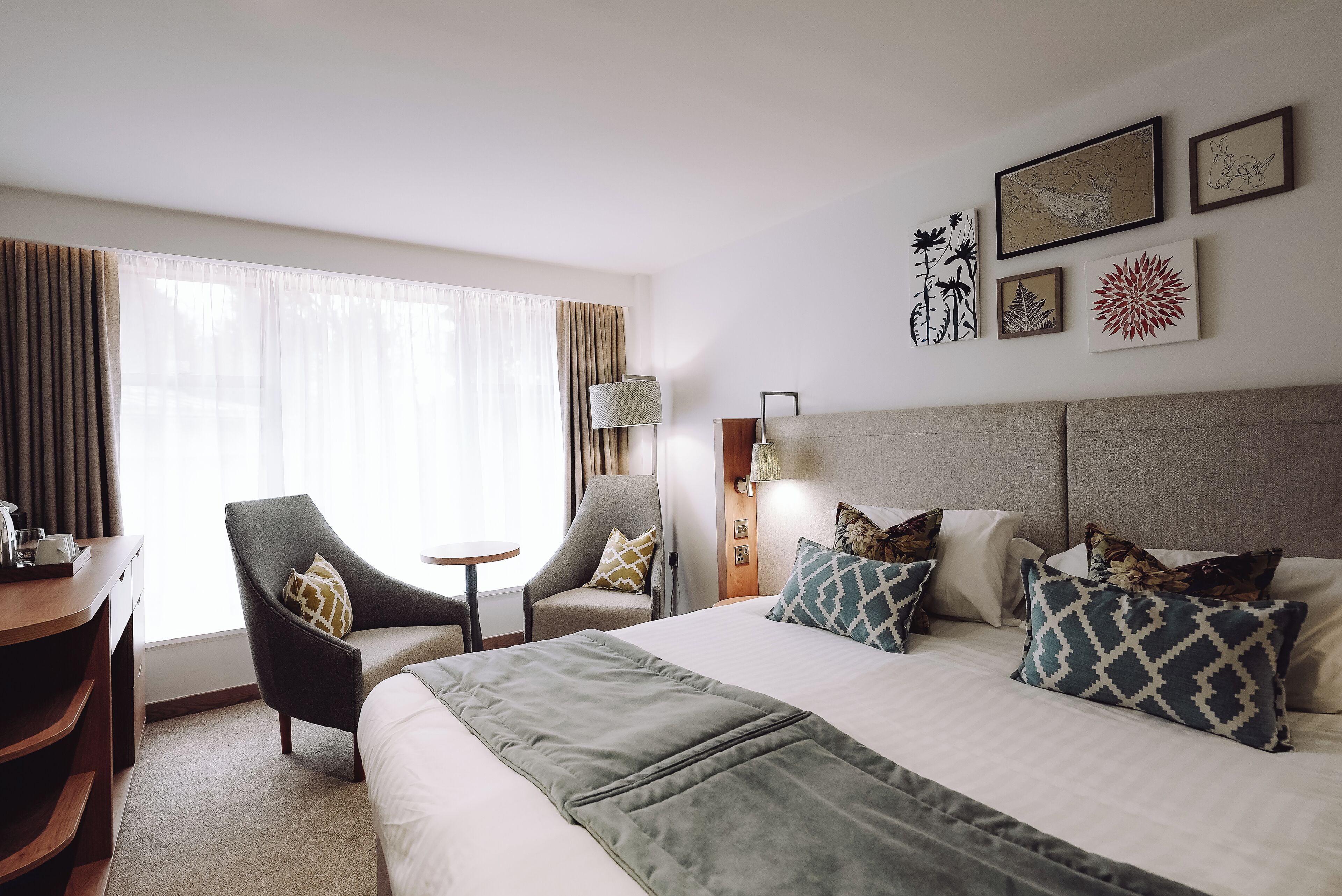Una camera d'albergo con un letto grande, due poltrone, un tavolino e una scrivania. Alle pareti si notano quadri incorniciati e una lampada sulla scrivania.