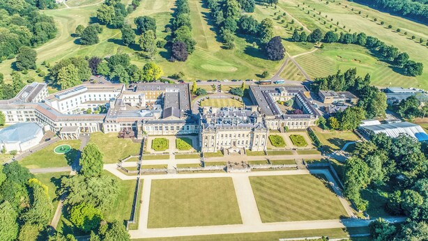 Una vista aerea di un ampio complesso architettonico storico con diverse ali, ampi giardini e un campo da golf sullo sfondo.