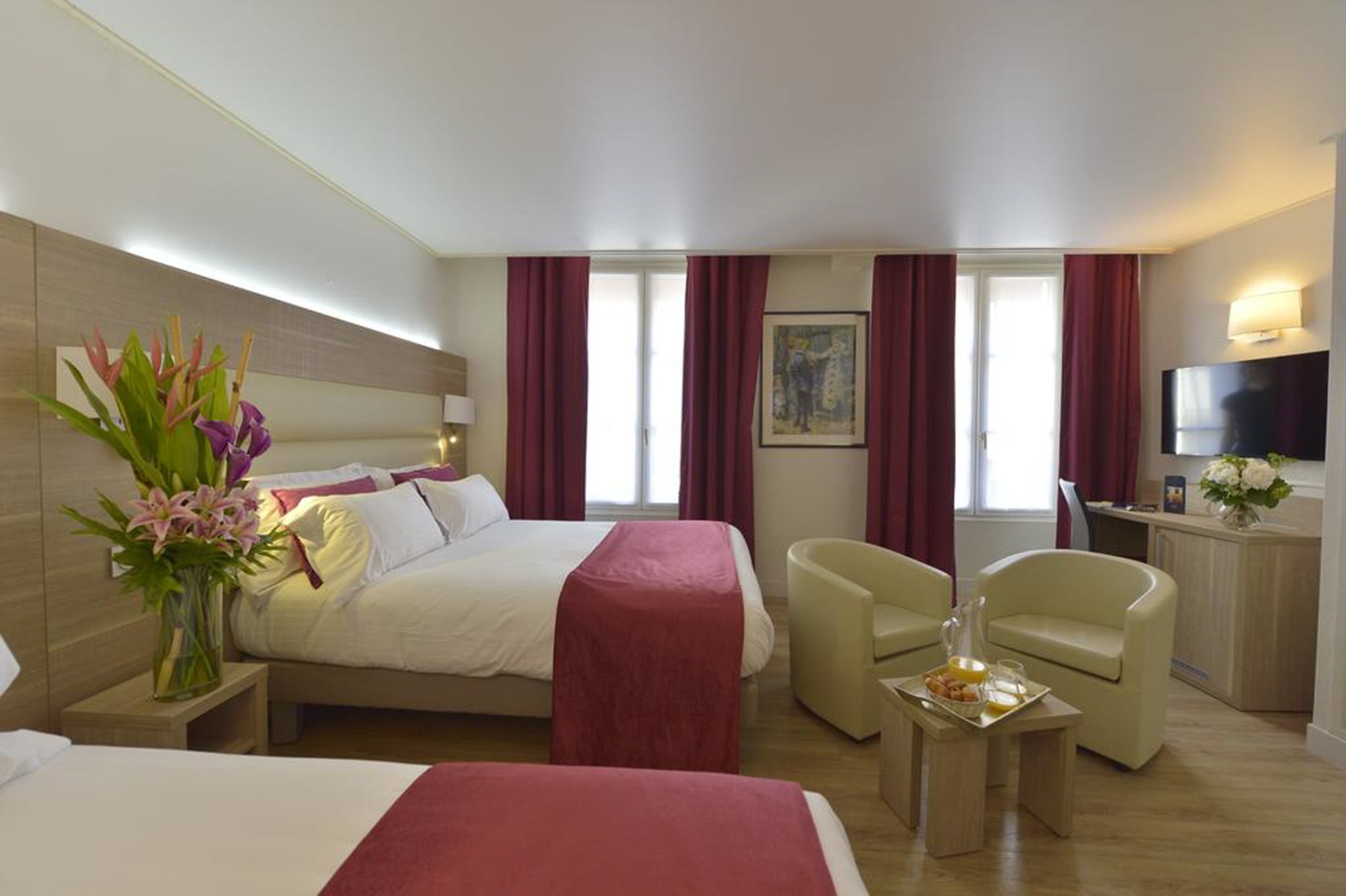 A hotel room with a bed, two armchairs, a small table with breakfast, a TV, and a painting on the wall.