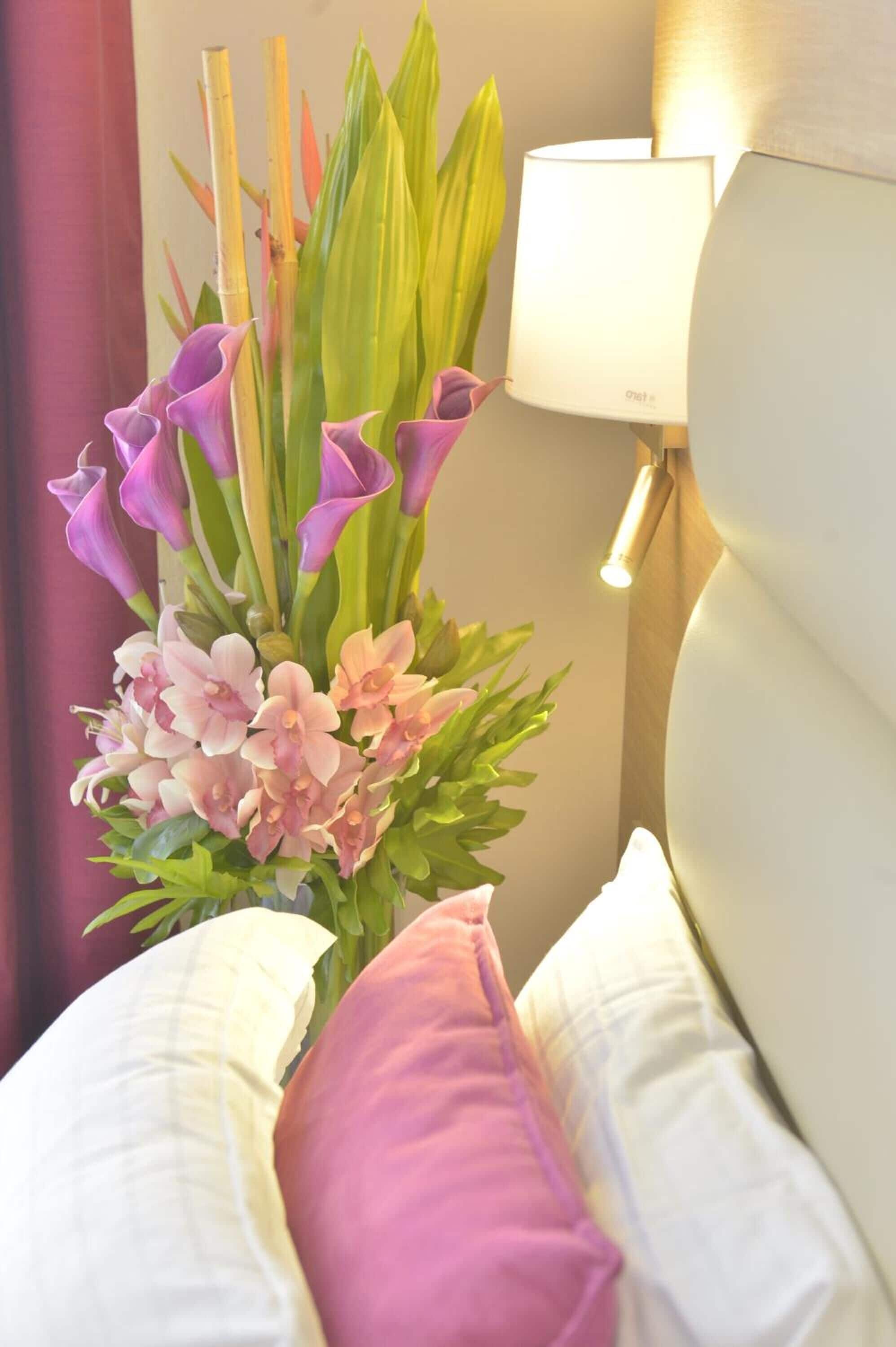 A neatly made bed with white and pink bedding, a decorative plant arrangement with purple and pink flowers, and a wall-mounted lamp.