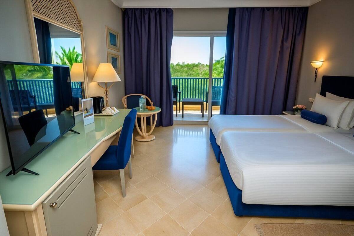 Habitación de hotel con cama, escritorio con televisor, silla, mesita de noche con lámpara y balcón con cortinas.
