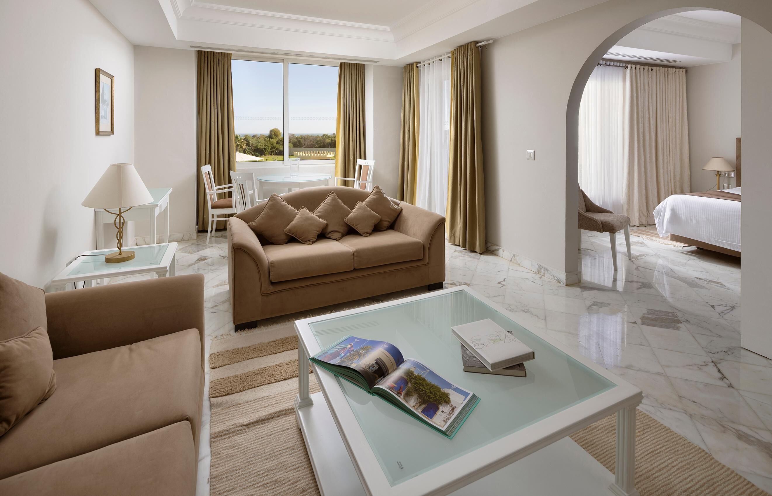 A modern hotel room with a sofa, a glass coffee table with magazines, a bed, and a view of the outdoors.