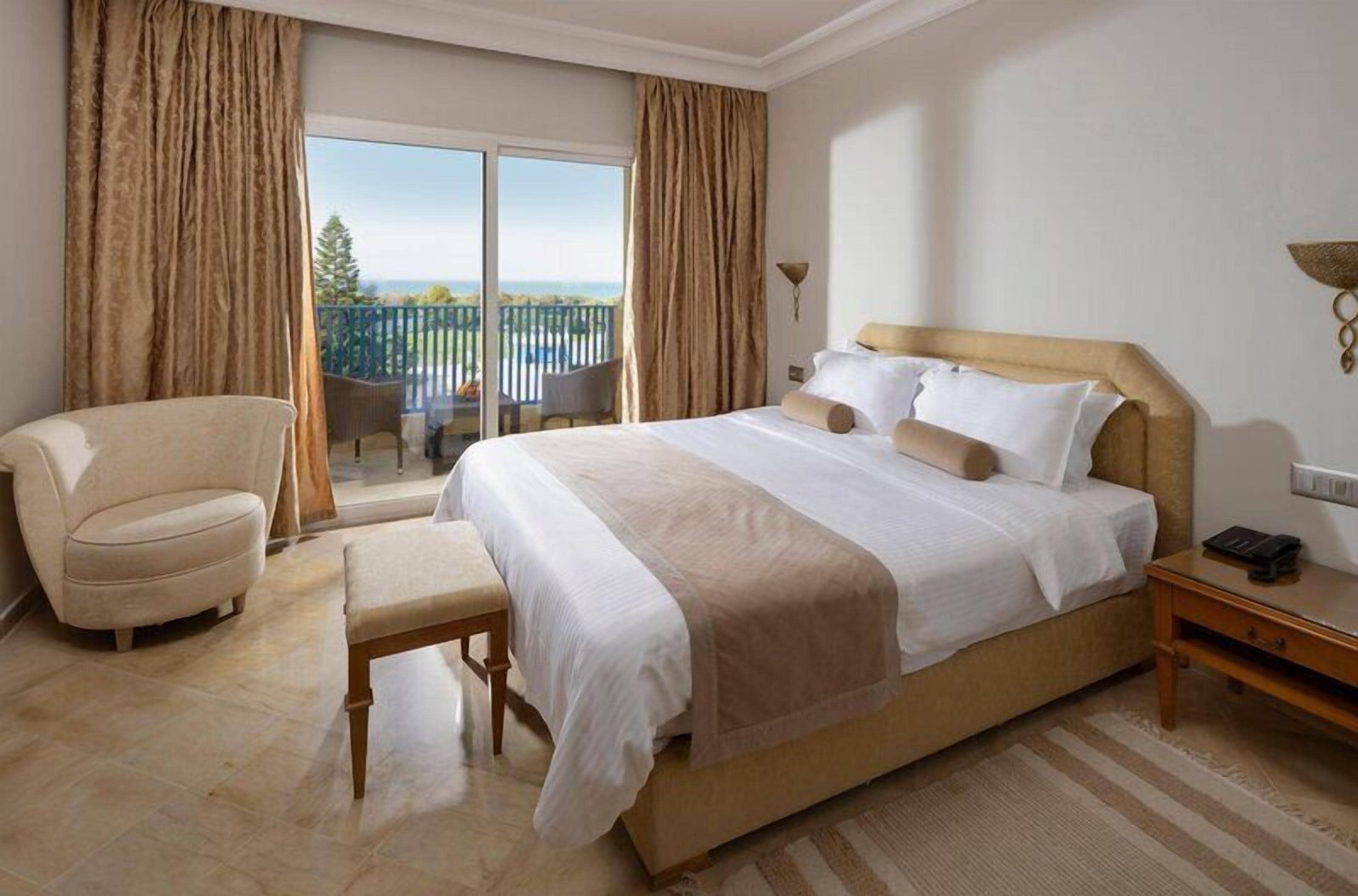 Quarto de hotel com uma cama grande, uma cadeira, um banco, uma mesa de cabeceira e vista para uma varanda com mobiliário.