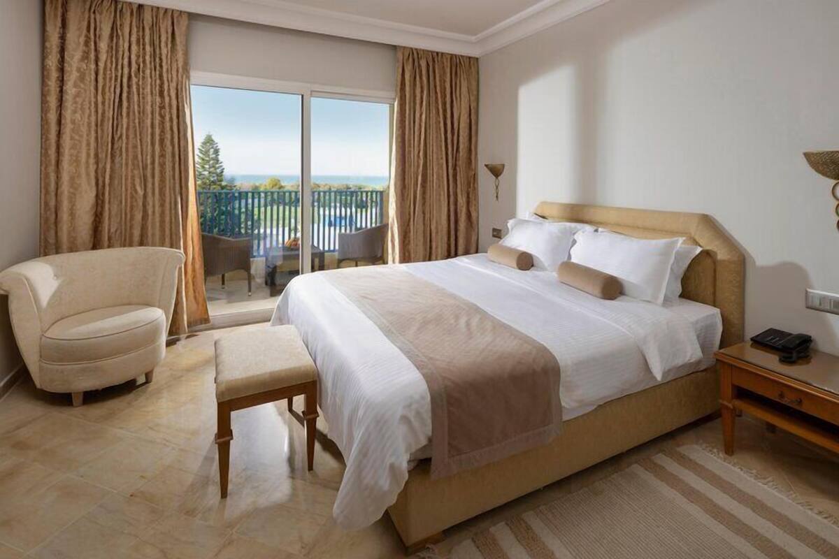 Habitación de hotel con una cama grande, una silla, un banco, una mesita de noche y vistas a un balcón con mobiliario exterior.