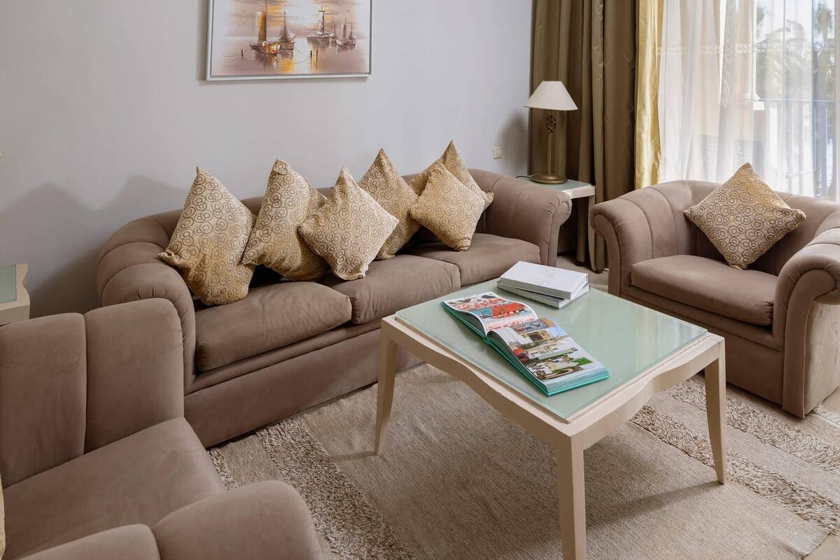 Una sala de estar con un sofá color beige, cojines decorativos estampados, una mesa de centro de vidrio y una obra de arte enmarcada en la pared.