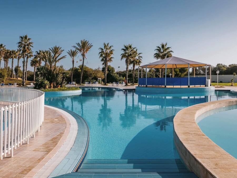 Una gran piscina curva con un pabellón y palmeras al fondo.