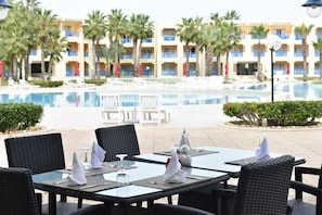 A poolside dining area with tables and chairs, a pool, and a hotel building with balconies and palm trees in the background. - Verdi Tunis Beach Resort (La Marsa)
