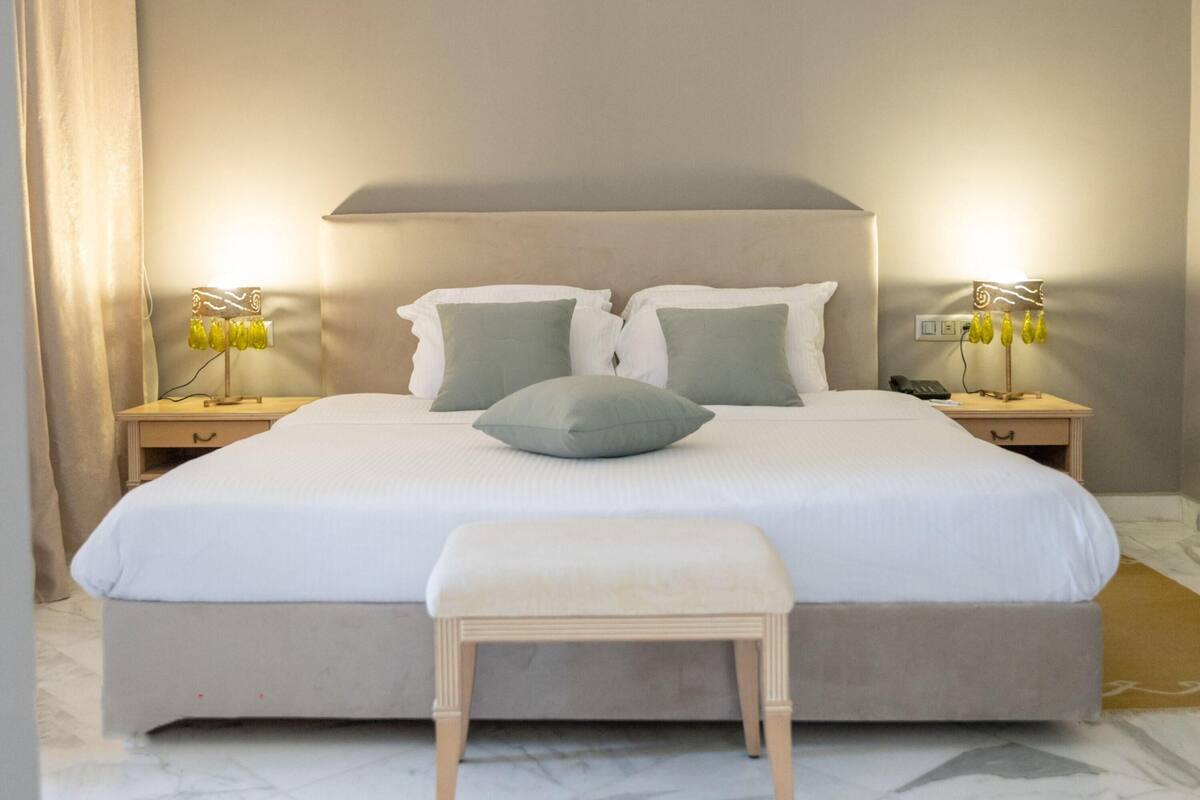 Una cama grande con cabecera, dos mesitas de noche con lámparas, y un banco frente a la cama.