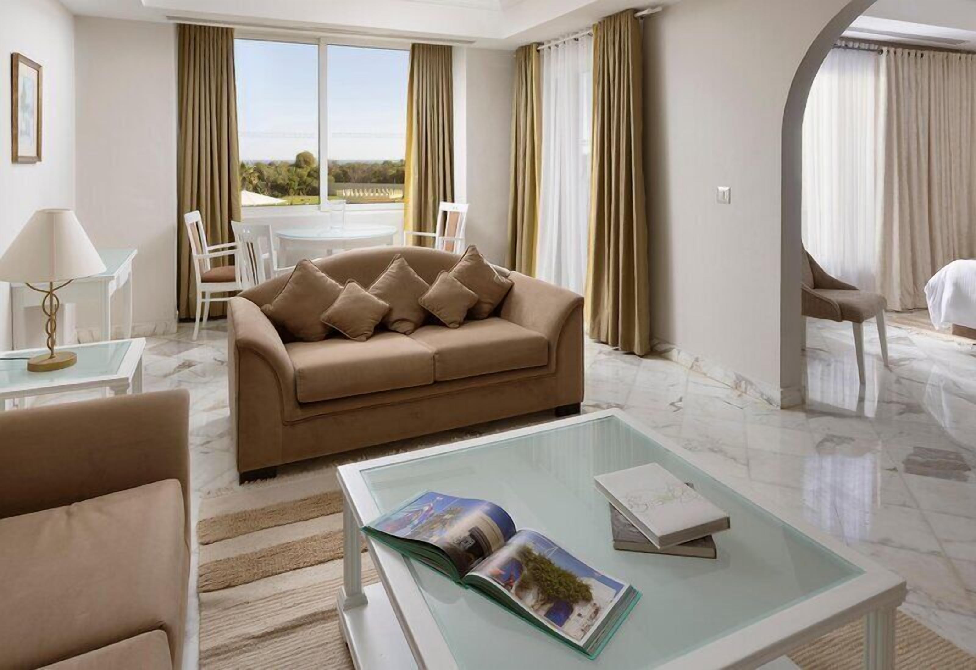 Una habitación de hotel moderna con un sofá color beige, una mesa de centro de vidrio y un área de comedor con una mesa y sillas blancas.