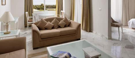 Una habitación de hotel moderna con un sofá color beige, una mesa de centro de vidrio y un área de comedor con una mesa y sillas blancas.