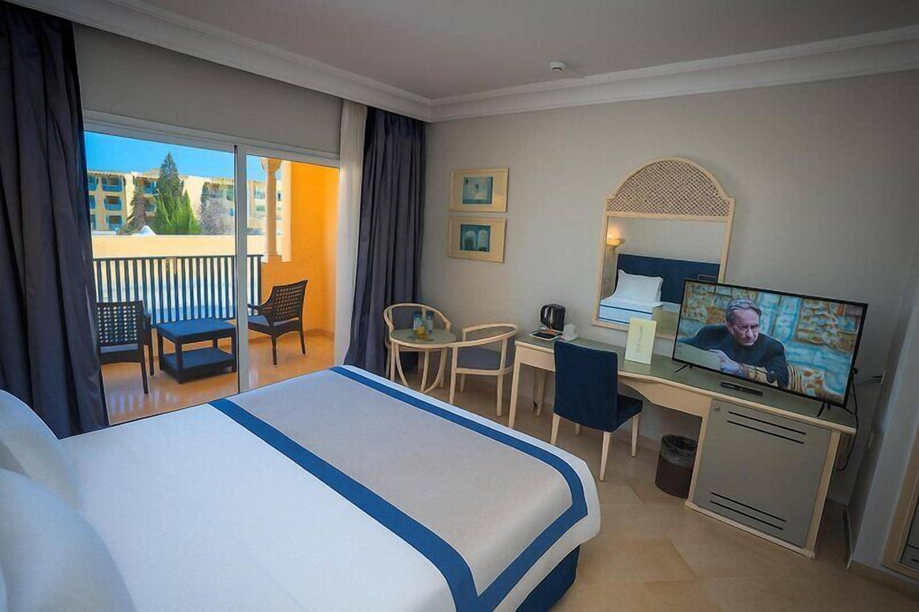 Habitación de hotel con cama, escritorio, televisor, silla y balcón con vistas a edificios.