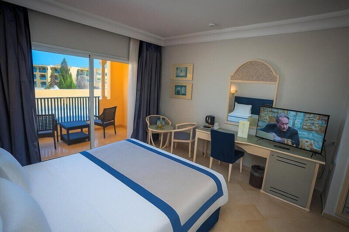 Une chambre d’hôtel comprenant un lit, un bureau, une télévision, une chaise et un balcon avec vue sur les bâtiments.