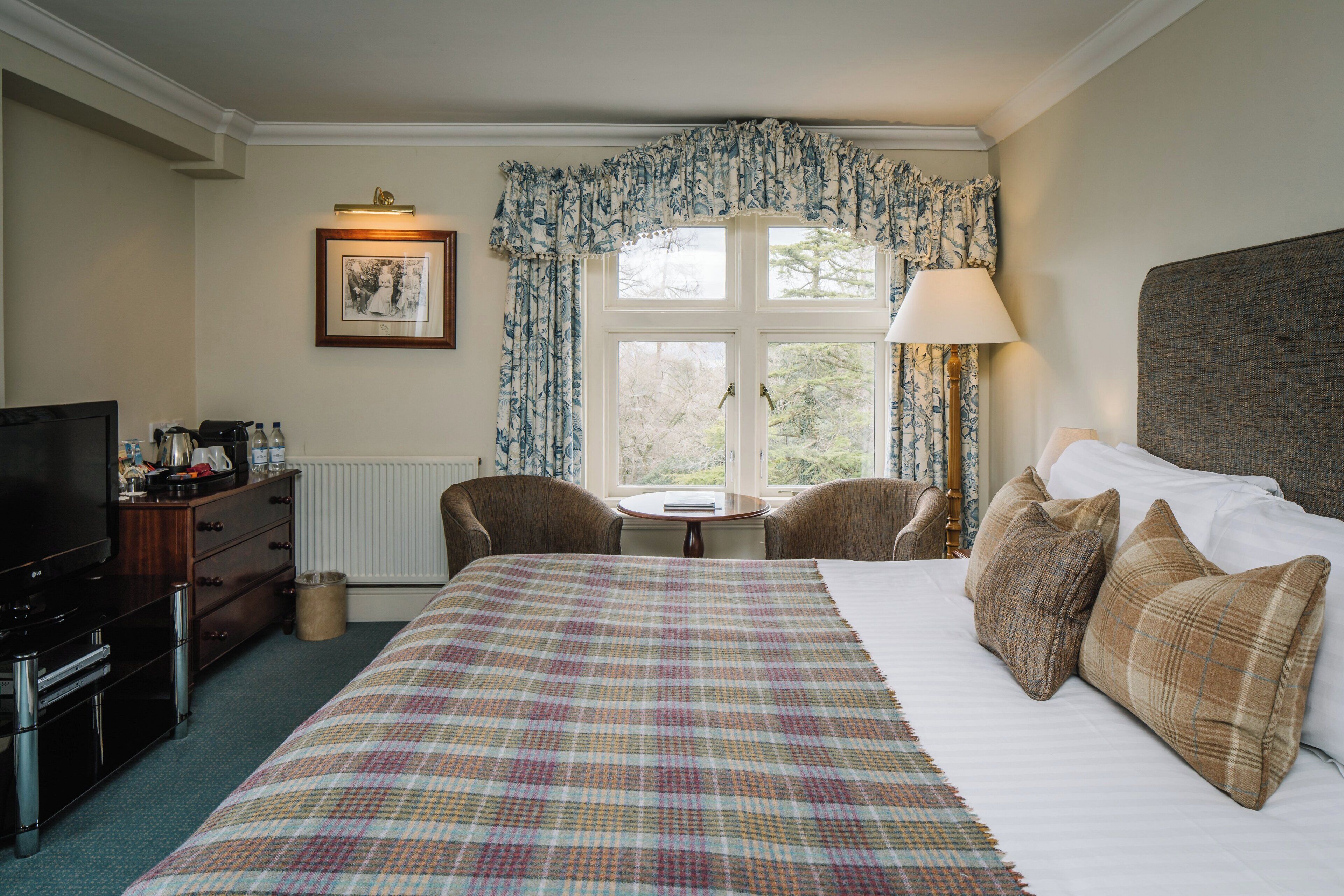 A hotel room with a large bed, a television, a small table, two chairs, and a window with curtains.