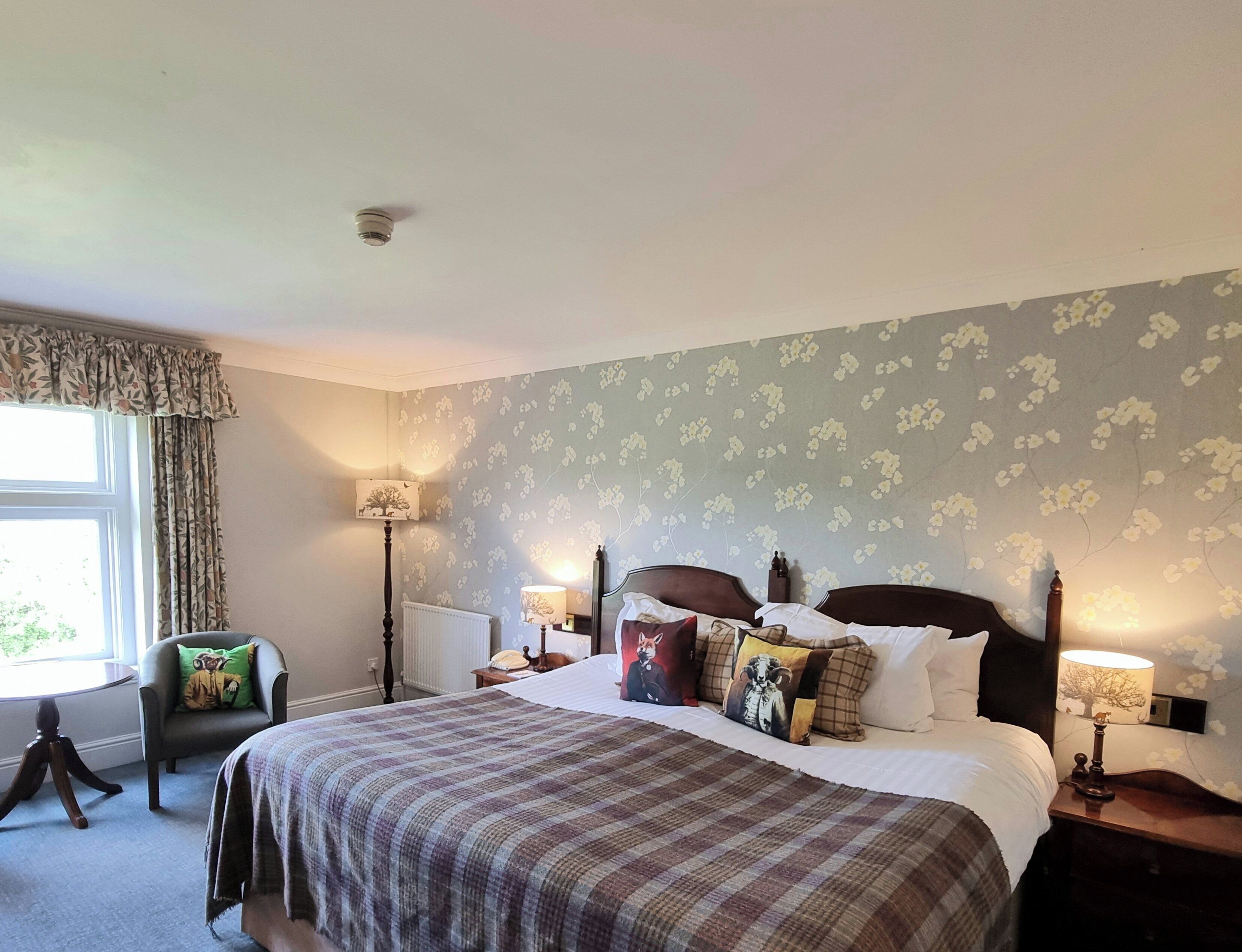 A neatly made bed with a plaid bedspread, flanked by two bedside tables with lamps, a chair with a cushion, and a window with curtains.