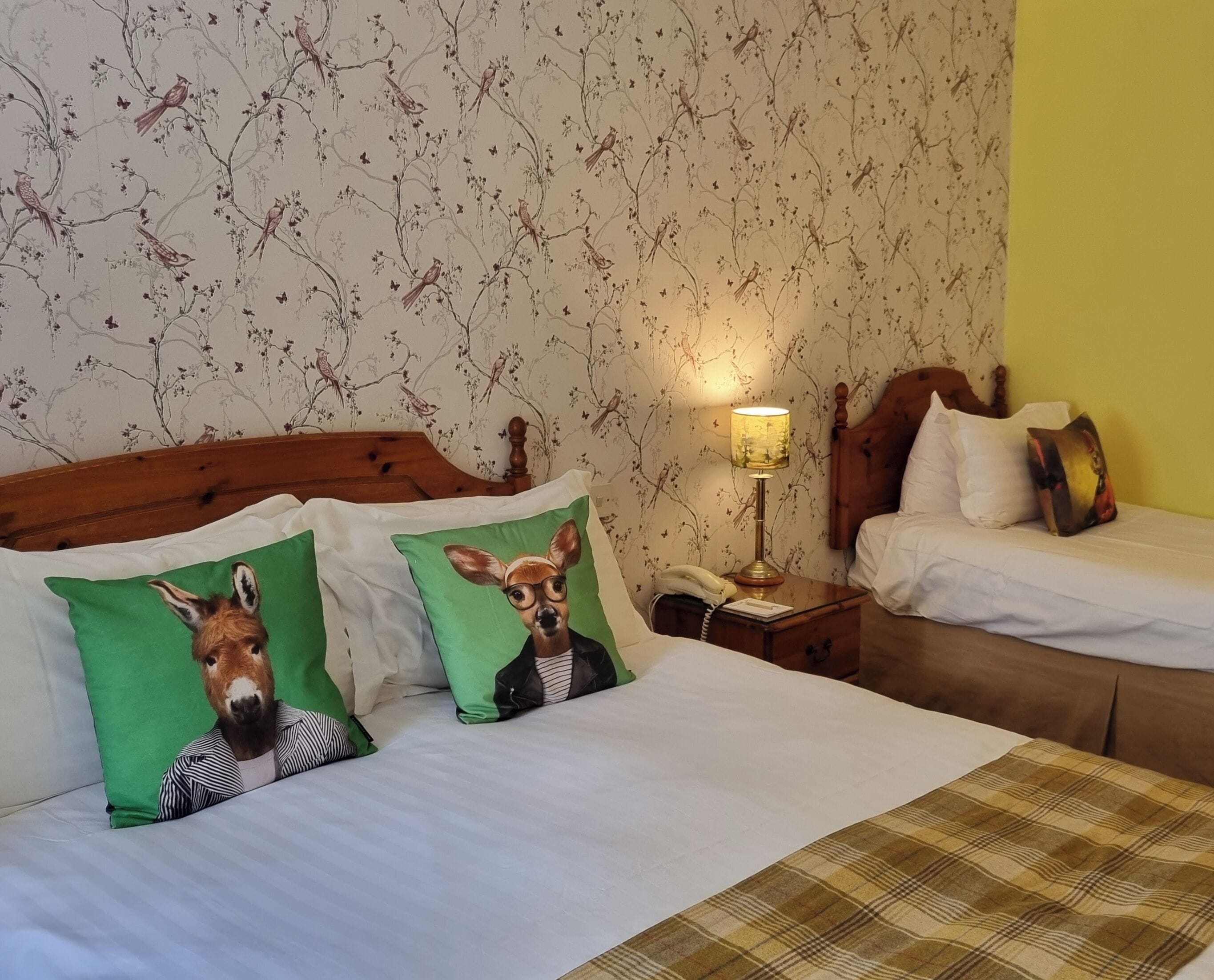 A hotel room with two beds, floral wallpaper, a wooden headboard, and a green and white pillow with a deer design.