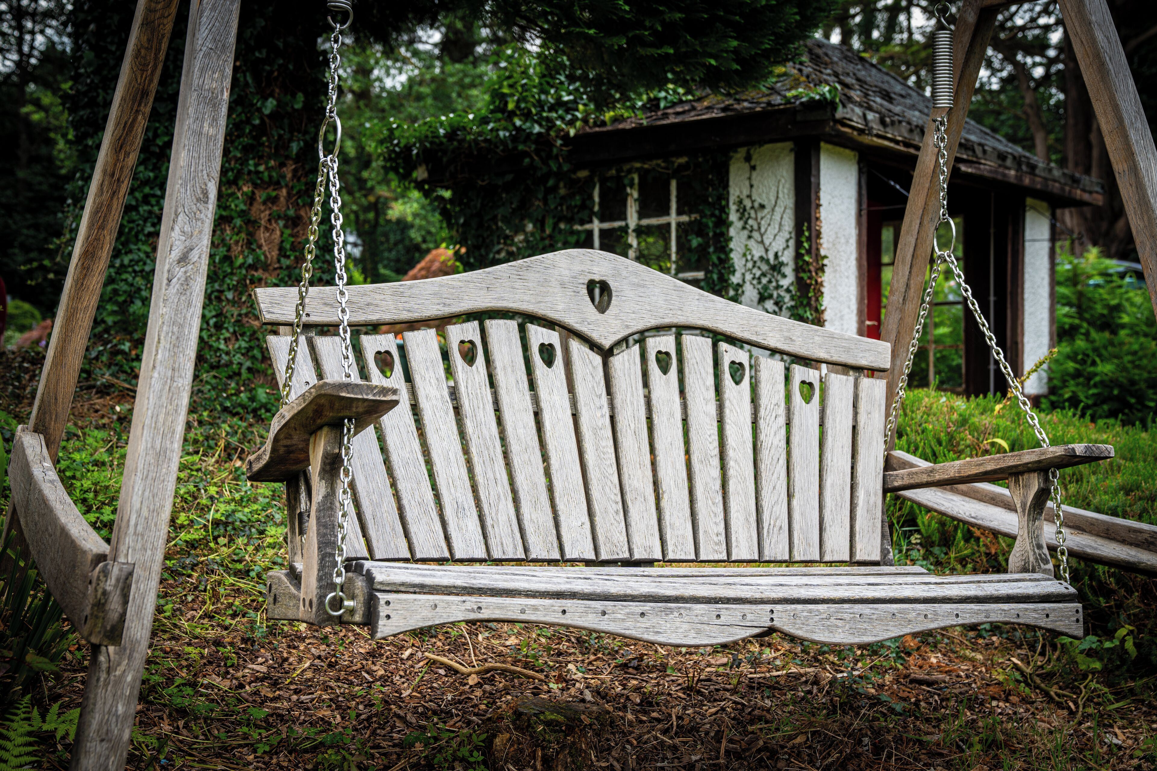 Un banc-balançoire en bois, orné de découpes en forme de cœur, installé dans un jardin avec une maison de style rustique en arrière-plan.