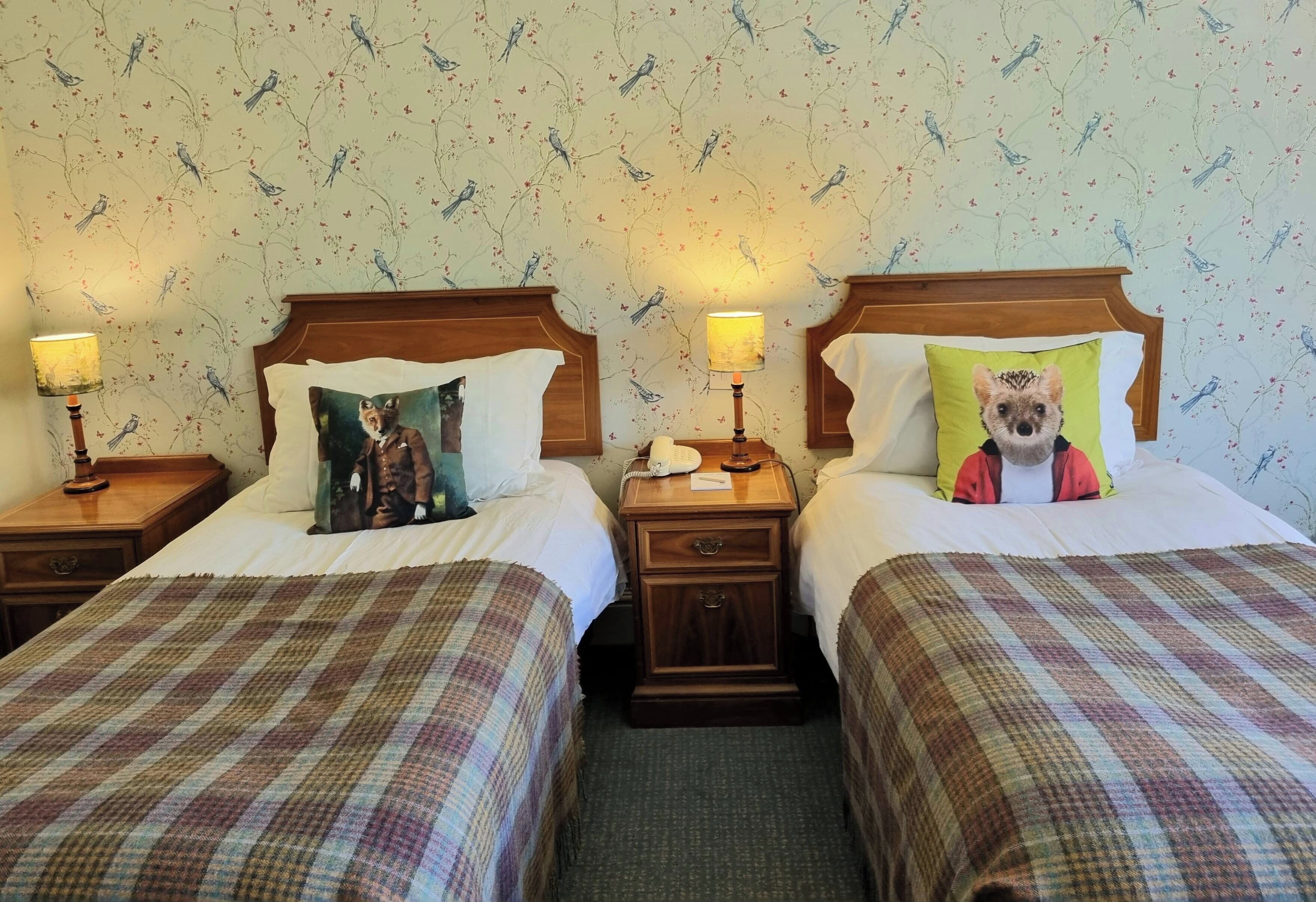 A hotel room with two beds, each with a wooden headboard and a nightstand. There are lamps on the nightstands, and the walls have a floral wallpaper.