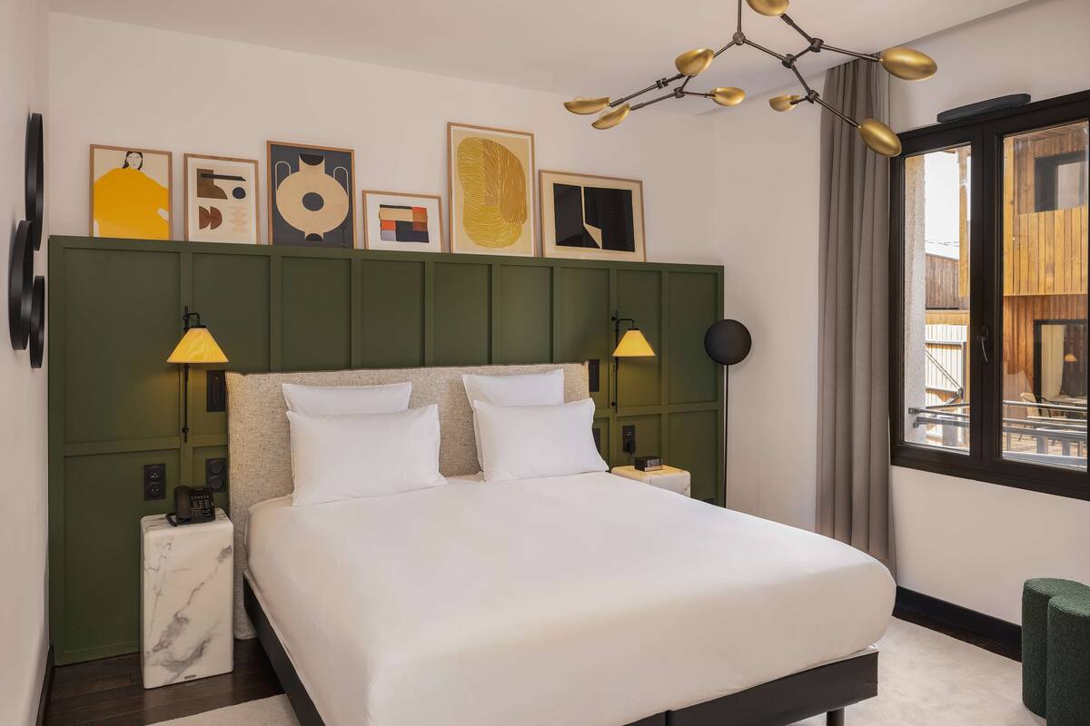 A modern hotel room with a large bed, two bedside lamps, a green headboard, and artwork on the wall.