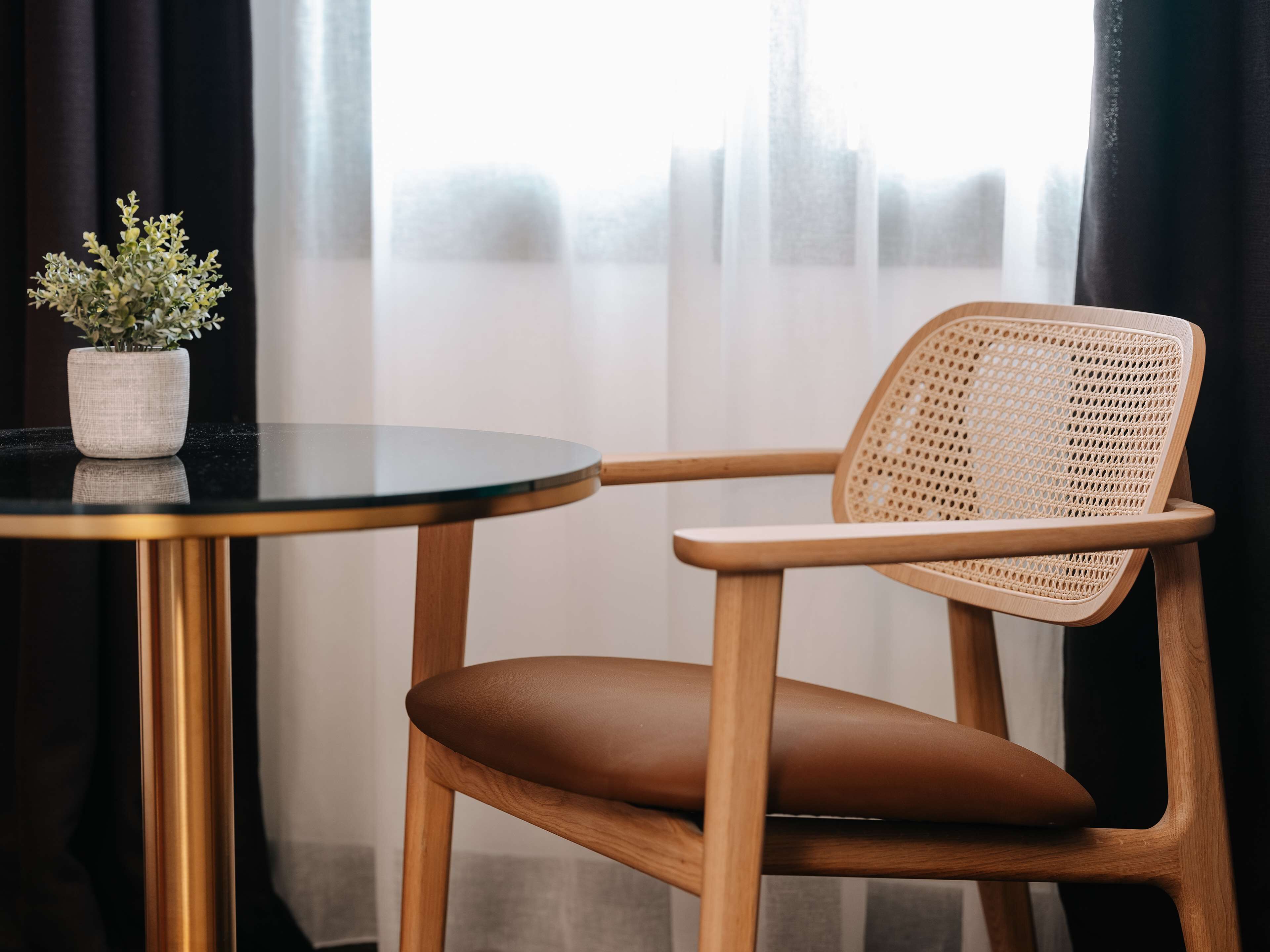 A round table with a potted plant, a wicker chair with a brown cushion, and a view of a building through sheer curtains.