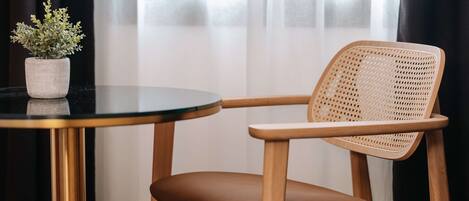 A round table with a potted plant, a wicker chair with a brown cushion, and a view of a building through sheer curtains.