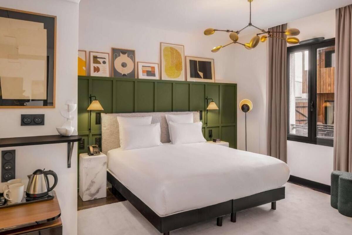 A hotel room with a large bed, bedside lamps, a green headboard, artwork on the wall, and a window with curtains.