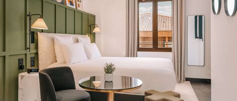 A modern hotel room with a bed, a round table with a plant, a chair, a green headboard, and artwork on the wall.