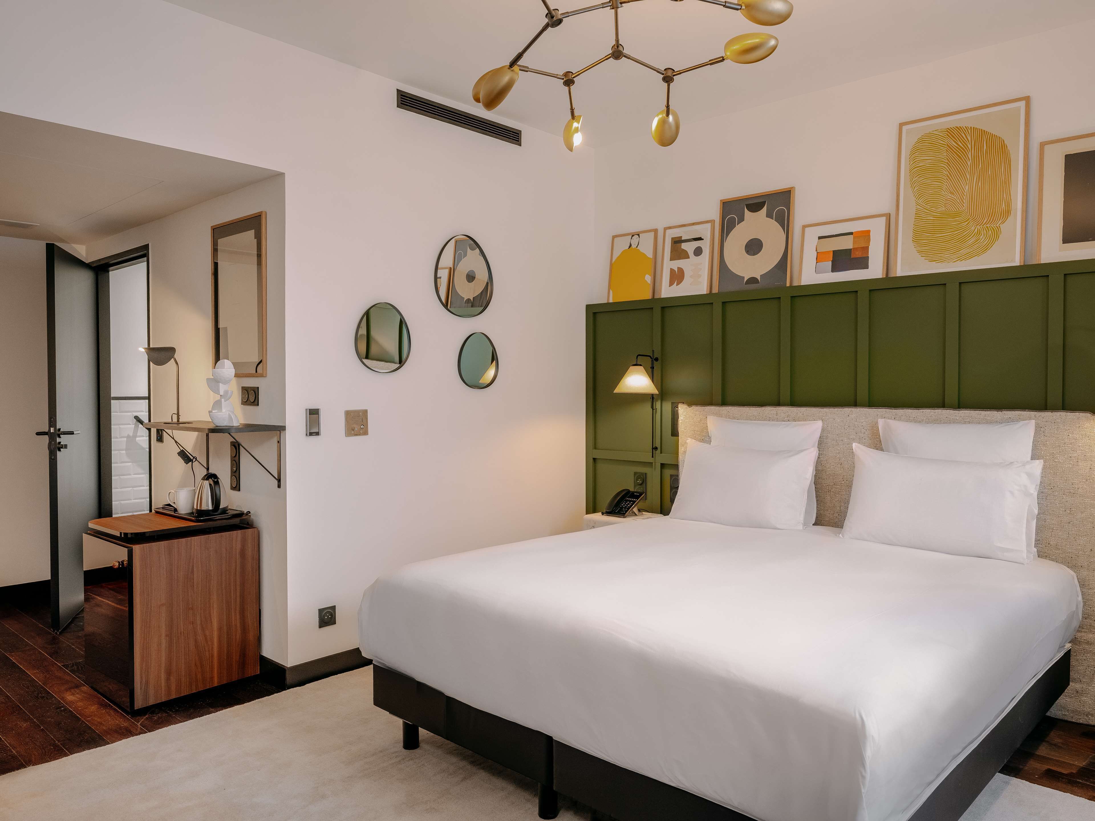 Ein modernes Hotelzimmer mit einem großen Bett, einem Schreibtisch mit Lampe, einem Spiegel und Kunstwerken an den Wänden.