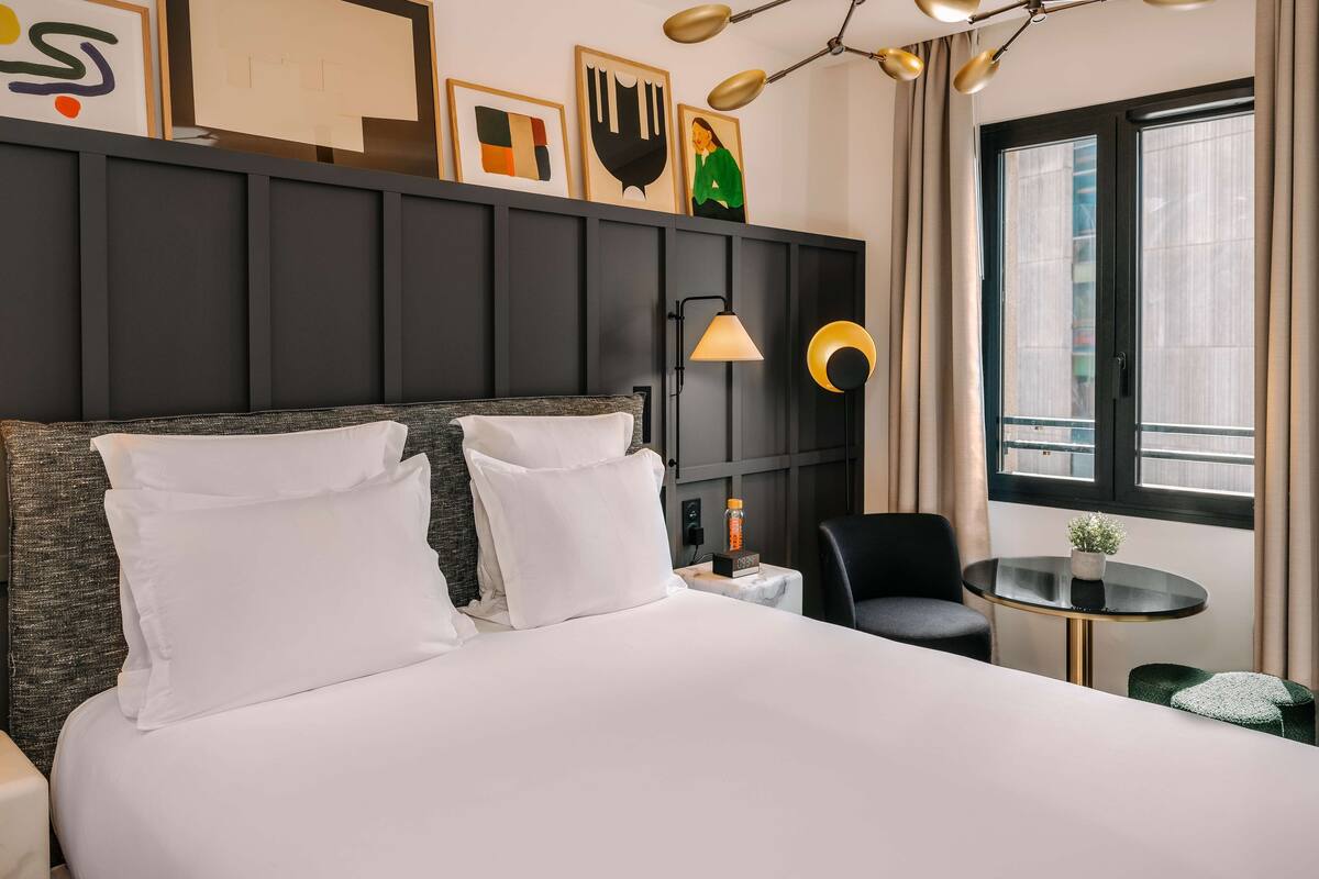A modern hotel room with a large bed, a desk, a chair, and a window with curtains.