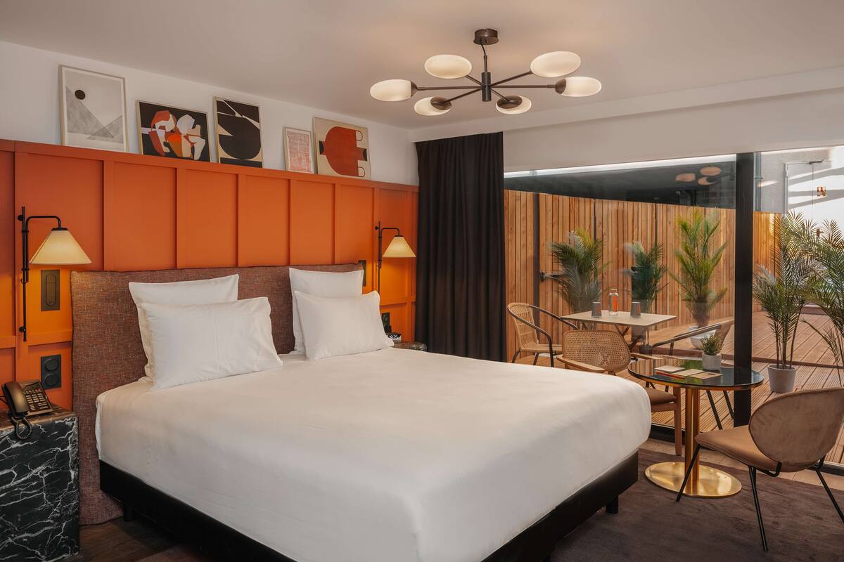 A hotel room with a large bed, two bedside lamps, a wooden headboard, a small table with chairs, and a view of the outdoors.